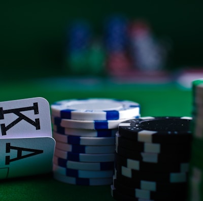 A casino scene featuring two playing cards, the ace and king of clubs, partially concealed by a player's hand. Nearby, there are stacks of multicolored poker chips on a green felt table, with blurred colorful background elements suggesting other chip stacks or cards.