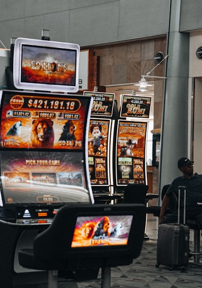 A collection of slot machines is situated inside a building with bright, colorful screens displaying various games and gaming themes. A person is seated nearby, seemingly relaxed with luggage beside them, and the atmosphere is lively with electronic displays.